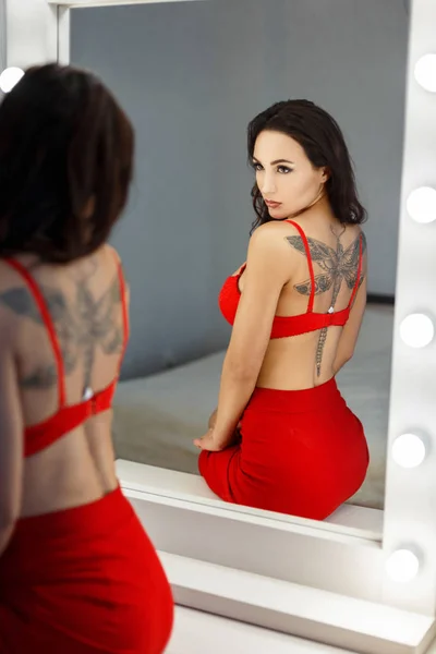Hermosa joven con un tatuaje en lencería roja sexy se ve en un espejo vintage con bombillas —  Fotos de Stock