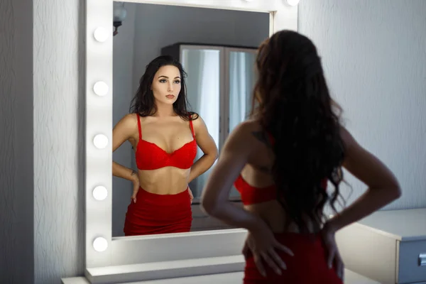 Belle jeune femme dans une robe rouge sexy avec un soutien-gorge se prépare pour une fête et regarde dans le miroir dans la chambre — Photo