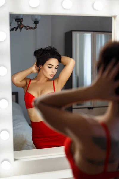 Beautiful sexy girl with a hairdo in red lingerie and a skirt looks in the mirror in the bedroom — Stock Photo, Image