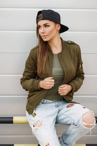 Mulher elegante bonita com sardas em um boné de beisebol em uma jaqueta verde na moda com jeans rasgados perto de uma parede de prata — Fotografia de Stock