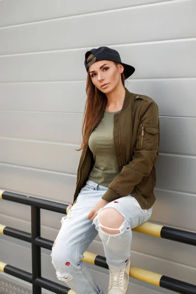 Menina bonita nova com sardas em um boné de beisebol em um estilo de rua em uma jaqueta verde militar na moda e calça rasgada azul senta-se perto de uma parede de metal — Fotografia de Stock