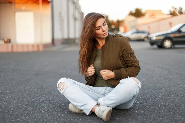 Mulher elegante bonita com cabelo em uma jaqueta na moda e jeans elegante rasgado senta-se no asfalto — Fotografia de Stock