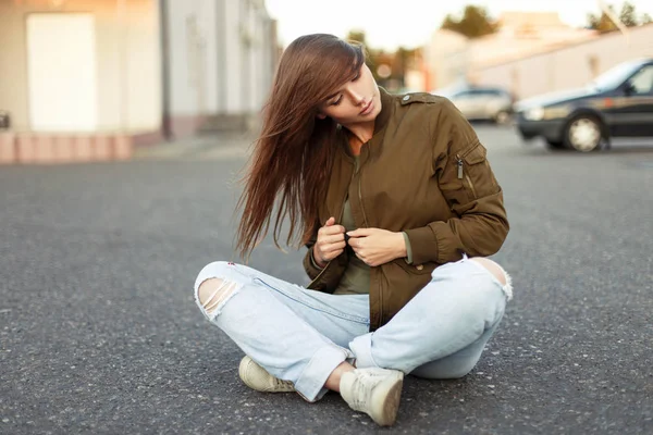 Mooie jonge vrouw met haar op gezicht in modieuze militaire jas en jeans zit op de weg — Stockfoto