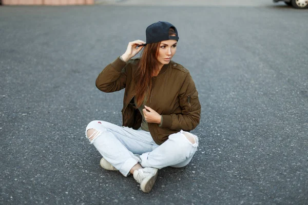 Beautiful stylish woman in a fashion military jacket with torn jeans sits on the asphalt — Stock Photo, Image