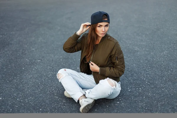 Vogue model woman in a baseball cap in a green stylish jacket and torn jeans sits on the asphalt