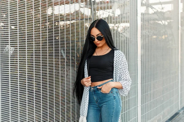 Modelo de mulher bonita com óculos de sol redondos vintage em um manto branco e jeans perto da parede — Fotografia de Stock