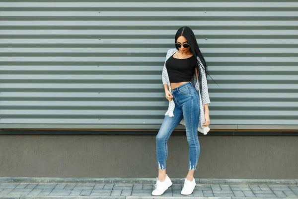 Красивая стильная женщина в модном белом плаще с черной футболкой и синими джинсами с высокой талией в белых туфлях возле металлической стены на улице — стоковое фото