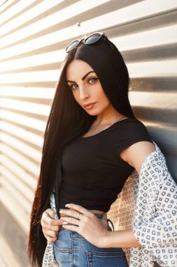 Güzel model ve kadın ile beyaz bir pelerin uzun saçlı sevimli bir yüzü güneşli bir metal bir duvarının yakınında kot ile siyah T-shirt