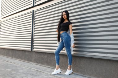 Güzel şık kadın beyaz ayakkabılarla moda giysiler içinde güneş gözlüğü ile modern bir duvara yakın duruyor