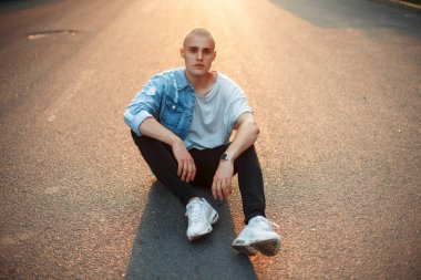Şık genç acımasız kel adam trendy denim giysiler içinde asfalt üzerinde günbatımında oturur.