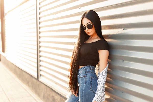 Güzel esmer kadın uzun saçlı beyaz pelerin ile modaya uygun güneş gözlüğü ve güneşli bir günde bir metal duvar yakınındaki yüksek bel ile kot pantolon siyah formaları