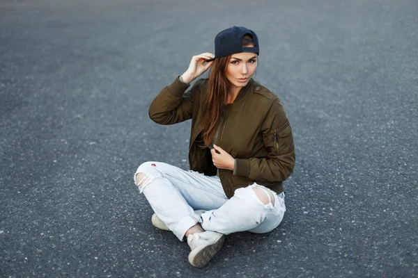 Vogue beysbol şapkası yeşil şık bir ceket ve yırtık kot modeli kadında asfalt üzerine oturur