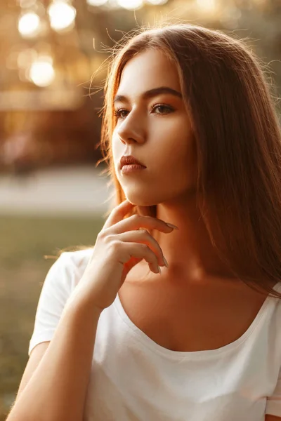 Retrato de moda de verão de uma jovem menina bonita em uma camiseta branca no parque ao pôr do sol — Fotografia de Stock