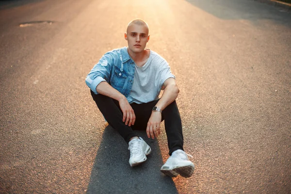 Şık genç acımasız kel adam trendy denim giysiler içinde asfalt üzerinde günbatımında oturur.