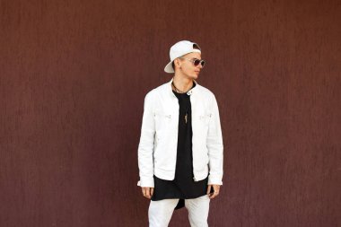 yakışıklı genç bir beyaz ceketli bir beyzbol şapkası, güneş gözlüğü ile duvara yakın duruyor