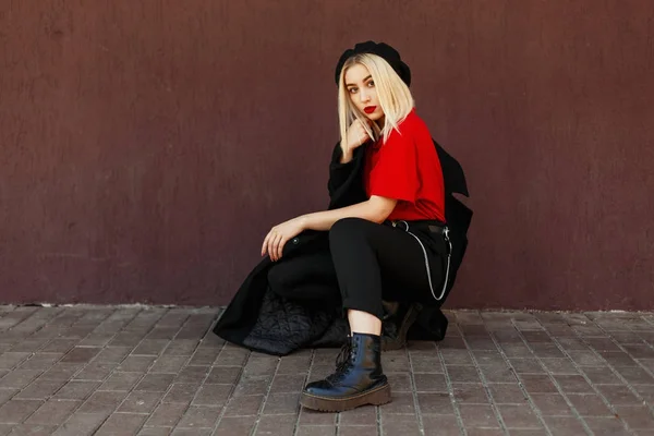 Modieuze mooie blonde vrouw met een zwarte baret met een zwarte herfst jas poseren op de straat in de buurt van de muur — Stockfoto