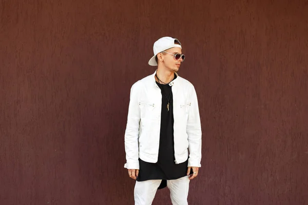Jovem bonito em uma jaqueta branca com um boné de beisebol em óculos de sol fica perto da parede — Fotografia de Stock