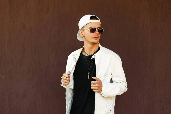 Hombre guapo en gafas de sol con estilo en una chaqueta de moda con una gorra de béisbol cerca de la pared — Foto de Stock