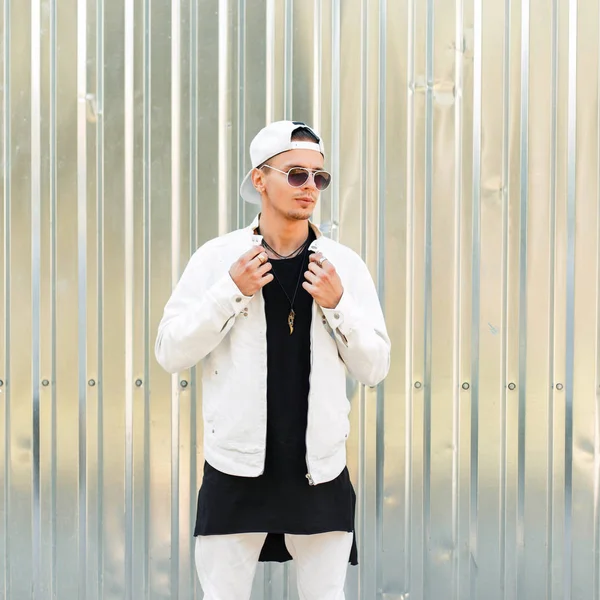 Hombre guapo de moda con gafas de sol en una chaqueta blanca con una gorra de béisbol cerca de la pared de metal — Foto de Stock