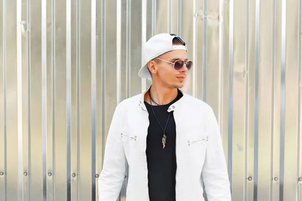 Stilig ung man i en fashionabel jacka med en vit keps i solglasögon den metall vägg — Stockfoto