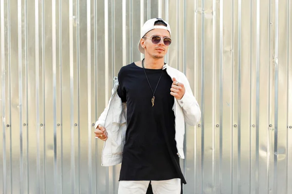 Homem elegante e bonito em uma camiseta preta com uma jaqueta branca em óculos de sol de moda perto da parede de metal — Fotografia de Stock