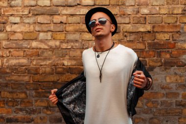 Yakışıklı genç bir siyah şapka ve siyah deri ceket tuğla duvara yakın poz güneş gözlüklü