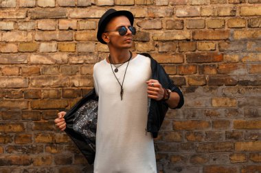 Yakışıklı genç bir şapka ve Beyaz tişört giyen güneş gözlüğü tuğla duvar yakınındaki bir siyah deri ceketli