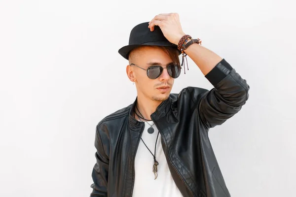Trendy knappe jonge man met hoed en zonnebril in een zwarte stijlvol jasje op een witte achtergrond — Stockfoto