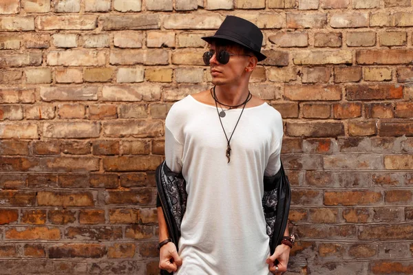 Handsome fashionable man in a white t-shirt, black leather jacket and hat with sunglasses posing near old brick wall — Stock Photo, Image