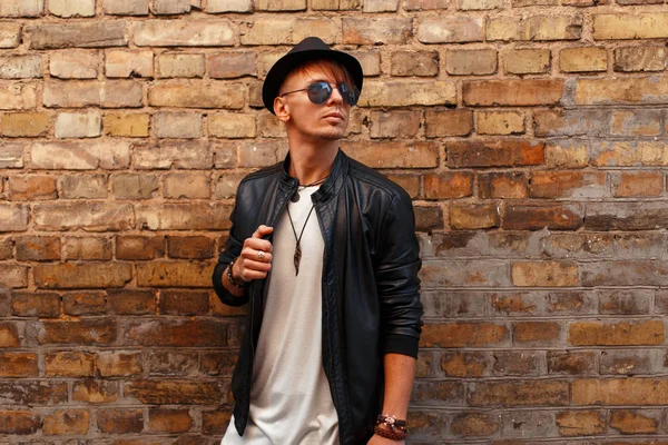 Handsome fashionable man with sunglasses in a fashionable hat in a leather black jacket near a brick wall — Stock Photo, Image