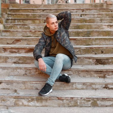 Merdivenlerde bir hoodie ve kot pantolon ile askeri ceket şık yakışıklı adam oturur