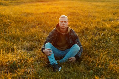 Kapüşonlu bir çim alanda günbatımında oturan bir kap ile şık bir askeri ceket şık yakışıklı adam