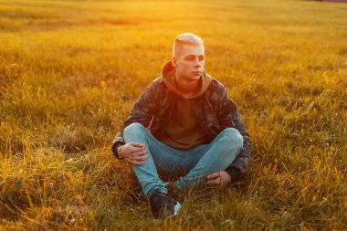 Gün batımında çimlere oturup kot ile askeri ceket şık yakışıklı adam