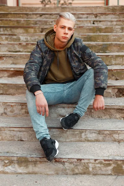 Joven con estilo en una chaqueta militar con una sudadera con capucha y jeans posando en las escaleras — Foto de Stock