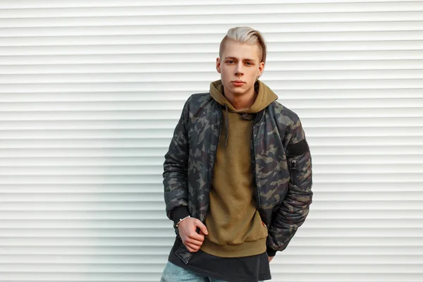 Guapo joven en chaqueta militar con estilo con una sudadera con capucha cerca de paredes de metal blanco — Foto de Stock