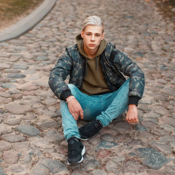 Красивий молодий чоловік у стильній військовій куртці з джинсами, що сидять на землі — стокове фото