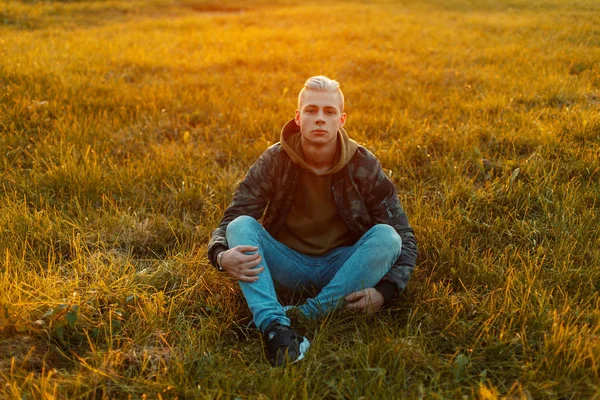 Kapüşonlu bir çim alanda günbatımında oturan bir kap ile şık bir askeri ceket şık yakışıklı adam