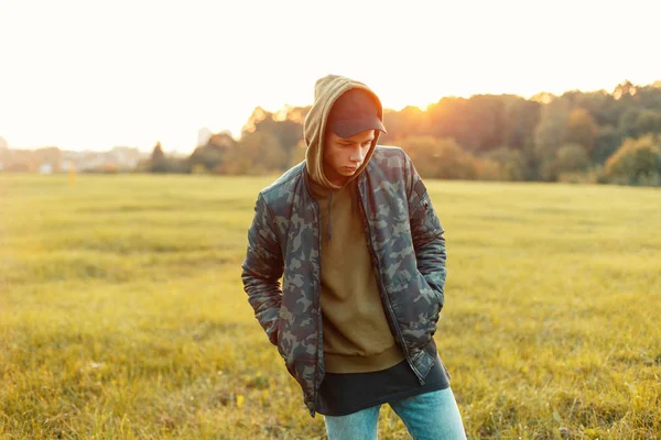 Bonito homem com um boné em uma jaqueta militar com um capuz na natureza ao pôr do sol — Fotografia de Stock