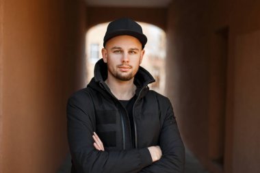 Yakışıklı şık genç hipster adam siyah beyzbol kap ve Kış moda ceket duruyor ve kameraya sokakta görünüyor