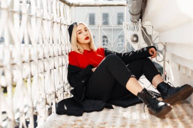 Bere ve güneşli bir günde dışarıda bot ile trendy siyah paltolu güzel genç şık kadın kırmızı dudaklar ile oturur