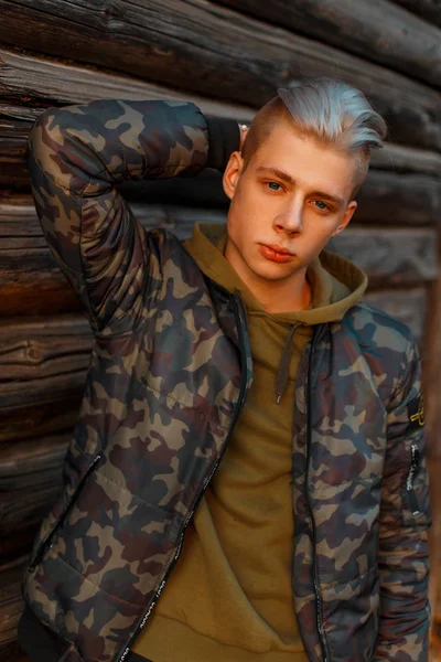 Handsome young man in a trendy military jacket with hoodie posing near wooden wall — Stock Photo, Image