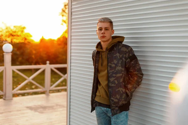 Стильный молодой человек в военной куртке с капюшоном, позирующий возле металлической стены на закате — стоковое фото