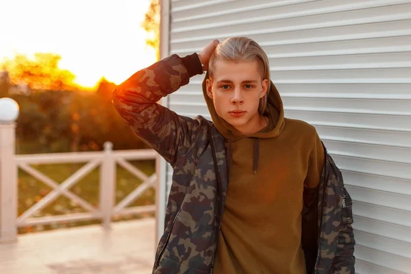 Guapo hombre de moda en una chaqueta militar con una capucha posando en la playa al atardecer — Foto de Stock