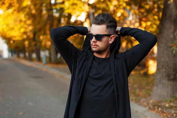 Man wearing sunglasses in a black hoody dresses a hood on the street, the autumn sunny day — Stock Photo, Image