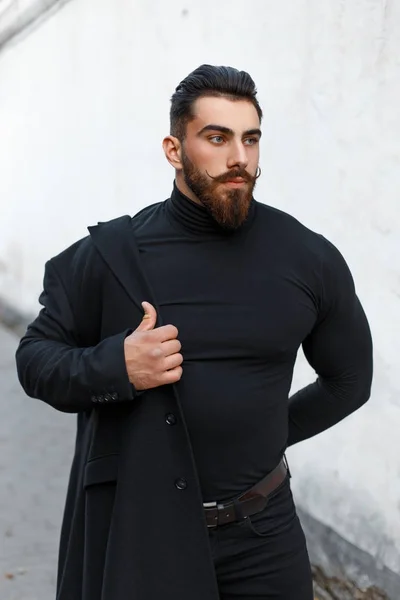 Módní pohledný muž s účes a plnovous klade na černý stylový kabát na ulici — Stock fotografie