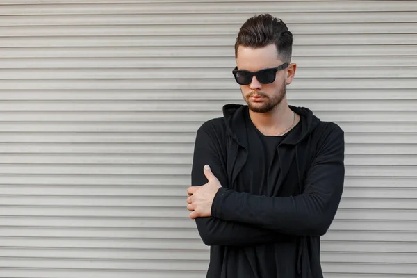 Stylish fashionable man in black sunglasses in a black hoody steals near a metal wall — Stock Photo, Image