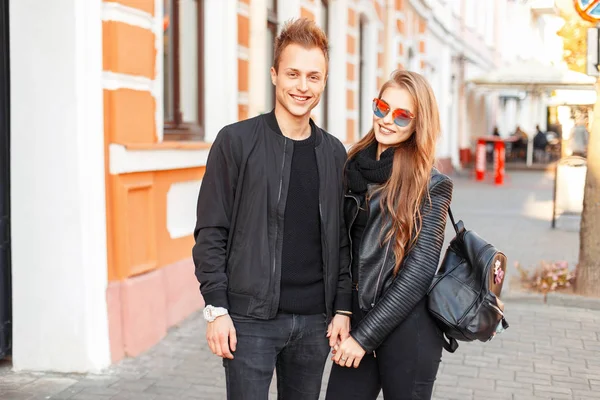 Stylish beautiful happy couple in sunglasses and fancy black clothes with a bag on the street — Stock Photo, Image