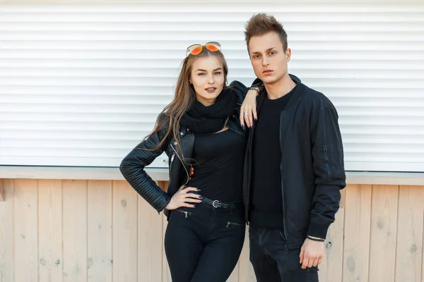 Hermosa pareja joven en chaquetas negras de moda vestido elegante de pie cerca de las paredes blancas de metal — Foto de Stock