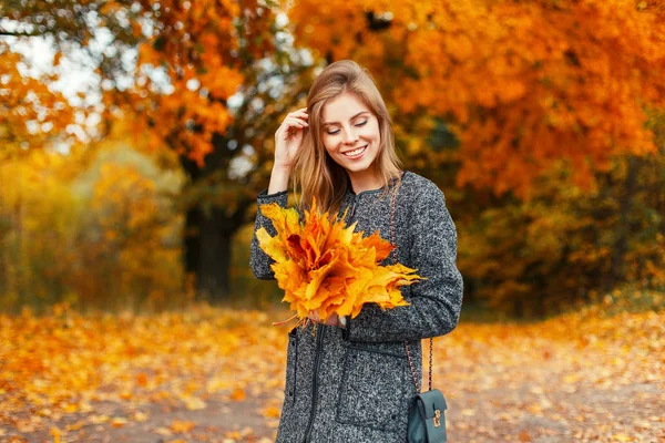 Beautiful happy woman with yellow autumn foliage in hands in trendy coat near colored trees — Stock Photo, Image