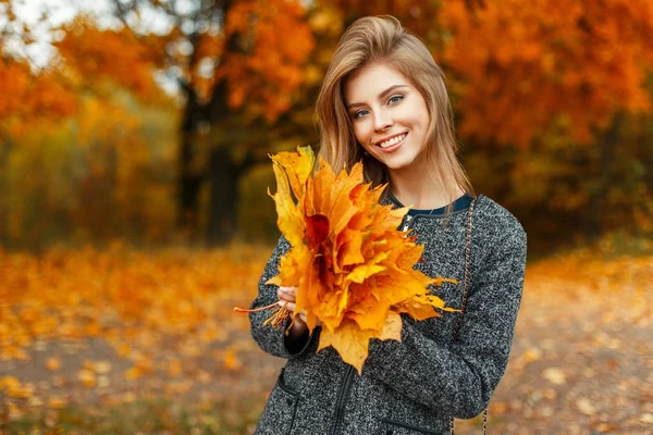 Glückliche Frau mit einem Lächeln im Mantel hält einen Strauß herbstgelber Blätter vor dem Hintergrund eines herbstlichen Parks — Stockfoto